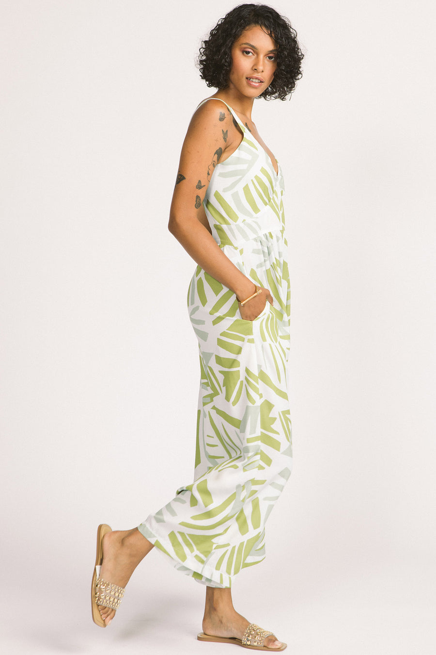 Side view of woman wearing green and white frond leaf print Zadie jumpsuit by Allison Wonderland. 
