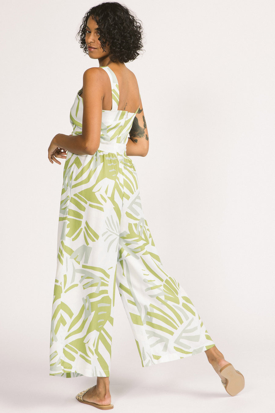 Back view of woman wearing green and white frond leaf print Zadie jumpsuit by Allison Wonderland. 